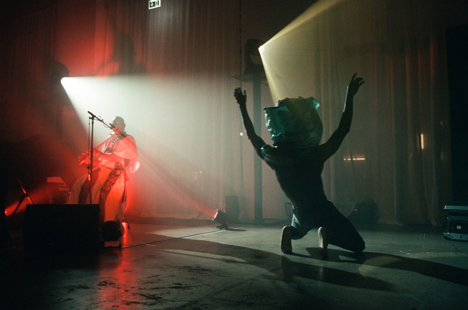 A dancer kneels with arms raised under a spotlight while a flutist plays in red light